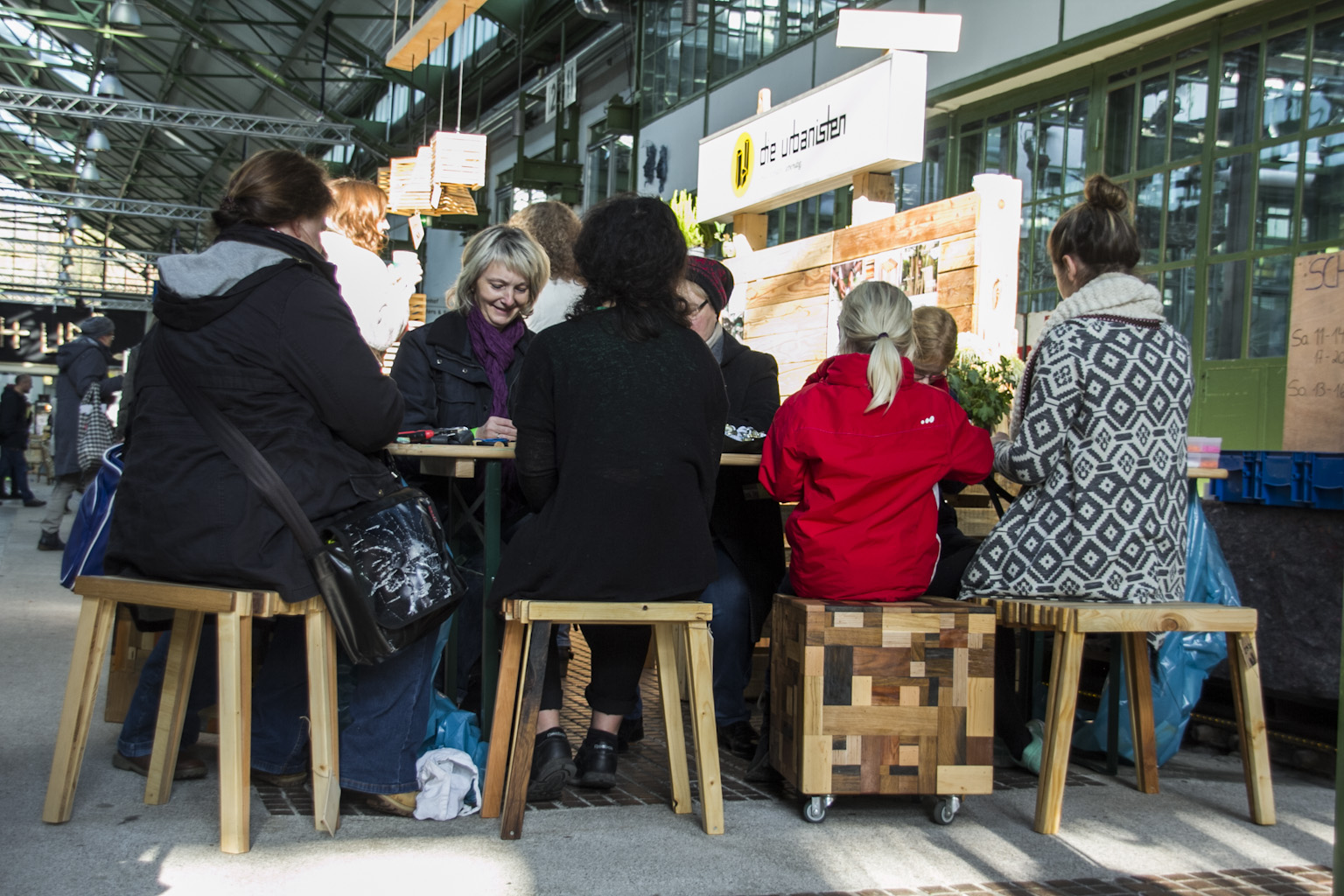 Eine Gruppe von Menschen sitzt an Holztischen in einer Markthalle und diskutiert miteinander.