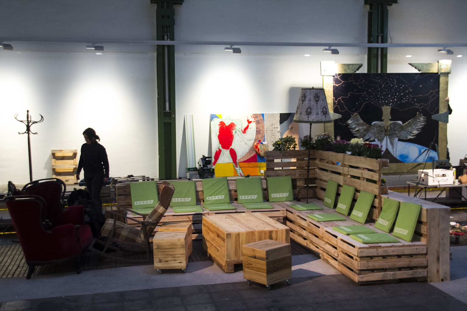 Ausstellung mit Palettenmöbeln, grünen Stühlen und bunten Kunstwerken in einer Industriehalle.